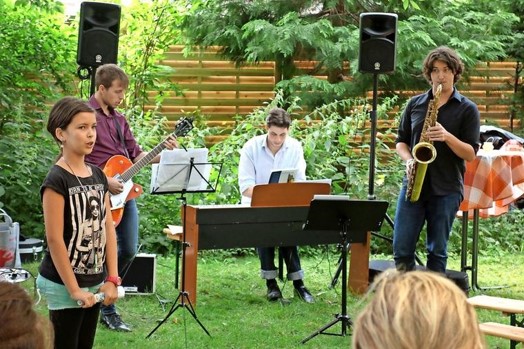 Vier Personen tragen draußen Musik vor. Eine Person sing, drei weitere spielen Gitarre, Klavier und Saxophon.