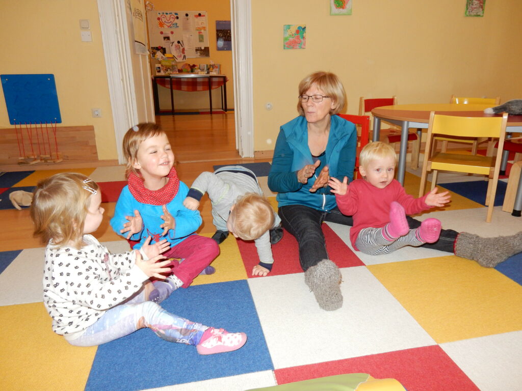 Vier Kinder und eine erwachsene Person sitzen klatschend auf einer bunten Matte in den Räumen des EKIDZ MiteinanderRaums