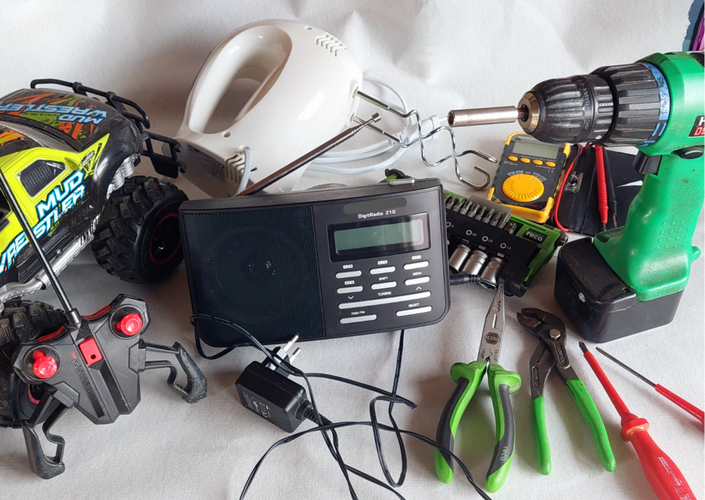 Verschiedene technische Geräte und Apparate, wie ein Mixer, ein Radio, ein ferngesteuertes Spielzeugauto liegen zusammengeworfen auf einer Stelle, daneben verschiedene Werkzeuge wie Schraubendreher, Zangen und ein Akkuschrauber